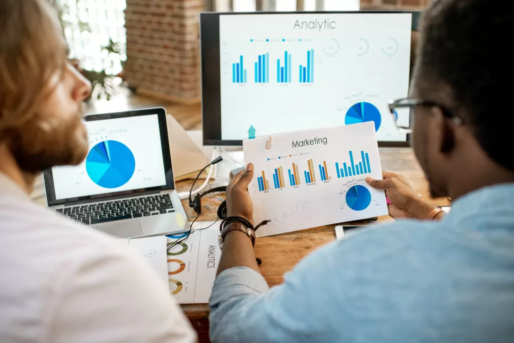 Two male coworkers working with analitycs sitting together at the working place with computer, laptop and documents. View from behind focused on the documents
